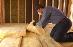 Technician kneeling on floor of unfinished space, wearing face mask, installing yellow batt fiberglass insulation.